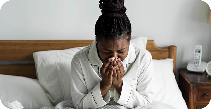 A woman sitting in bed in white pajamas, sneezing into a tissue.