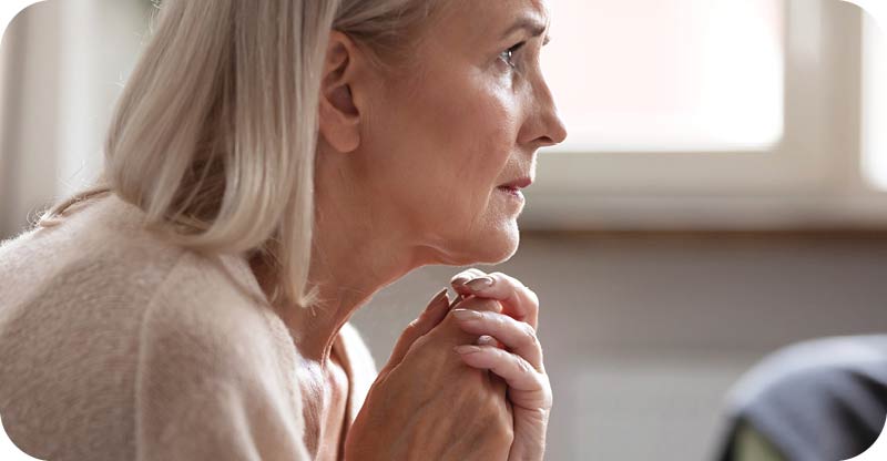 Side profile of a concerned senior woman with grey hair, resting her chin on her clasped hands while looking pensively out a window.