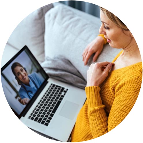 A woman in a yellow sweater sitting on a couch and participating in a telehealth video call with a female healthcare provider on her laptop.