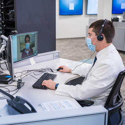 A healthcare professional in a white coat, wearing a face mask and a headset, conducting a telehealth consultation on a computer screen.