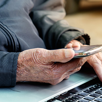 A close-up shot showing the weathered hands of an older adult holding a smartphone, with a laptop keyboard visible in the blurred background.