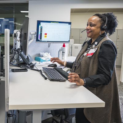 A healthcare professional wearing a headset and smiling while working at a standing desk with a computer monitor, illustrating a patient-driven triage or telehealth setting.