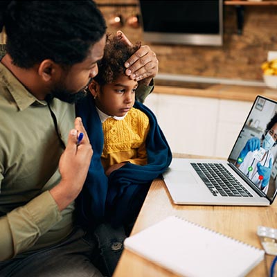 A father sitting at a kitchen table holding his young daughter, who is wrapped in a blue blanket, while participating in a telehealth video call with a doctor on a laptop.