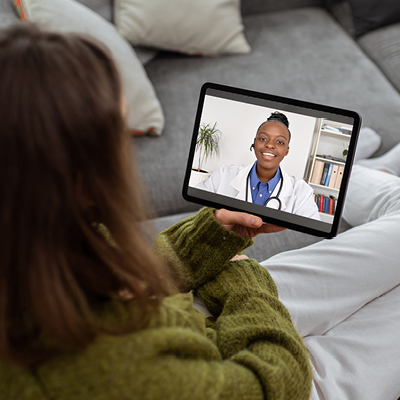 A person sitting on a couch and holding a tablet during a telehealth appointment with a female healthcare professional.