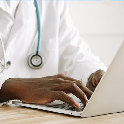 Close-up of a healthcare professional in a white lab coat with a stethoscope around their neck, typing on a laptop computer.
