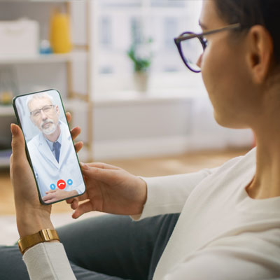 A person wearing glasses holds a smartphone during a virtual urgent care appointment with a male doctor on the screen.