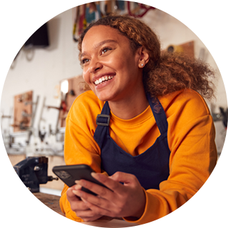 A smiling woman in a yellow shirt and blue apron holding a smartphone in a workshop setting.
