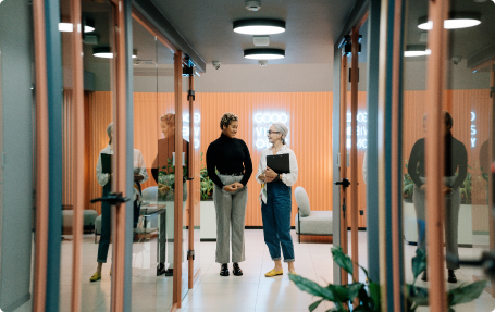 Two professional women standing in a modern office hallway with glass-walled meeting rooms and warm orange accents.