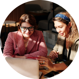 A woman in a pink blazer and glasses and a woman with a blue headband looking at a laptop screen together in a brightly lit office setting.
