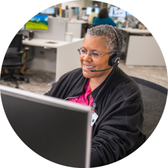 A female healthcare professional wearing a headset and pink scrubs, smiling while working at a computer in an office setting.