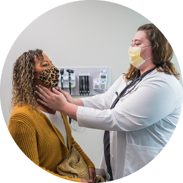 A healthcare provider wearing a mask examines a seated patient’s neck and throat in a medical exam room, both wearing face masks.